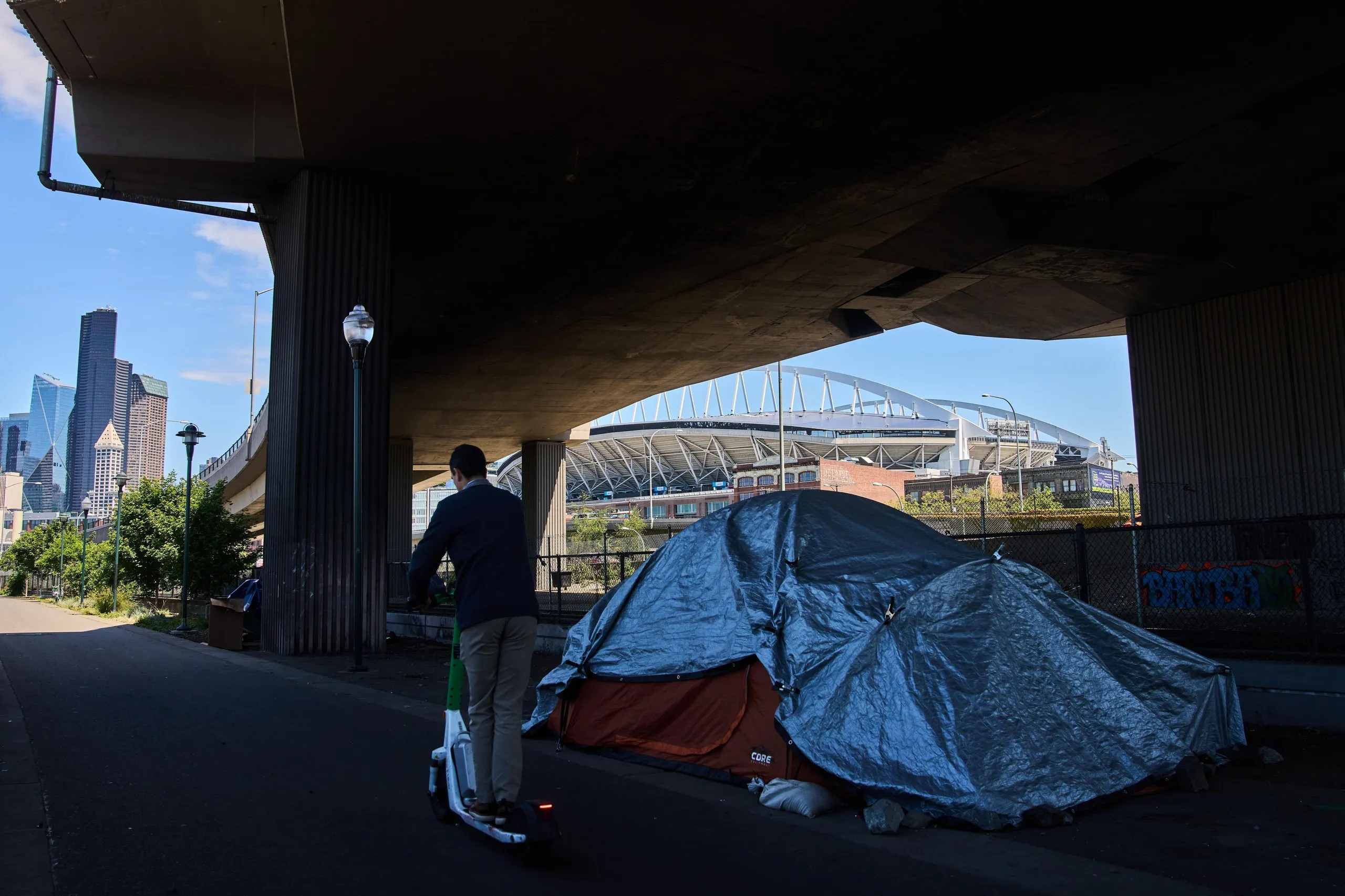 Seattle Stadium is seen in the background as a person rides a scooter past a series of tents on a trail near the stadium